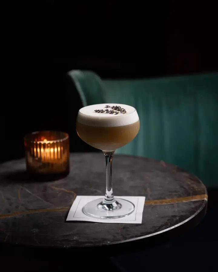 A lavender topped cocktail on a marble table by candlelight