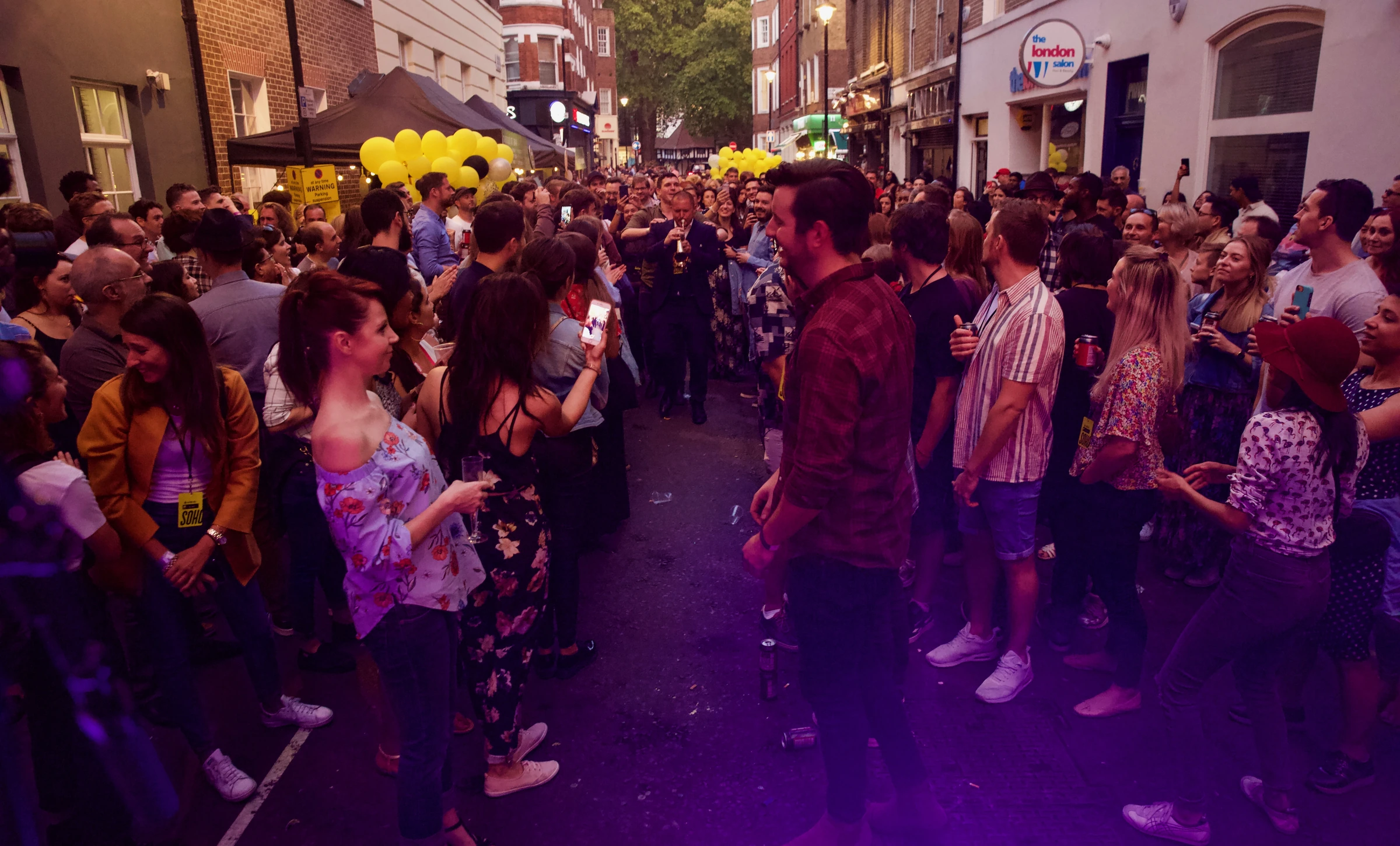 A trumpet player leads the way through a packed Soho street closed for a festival party, with yellow balloons along the street