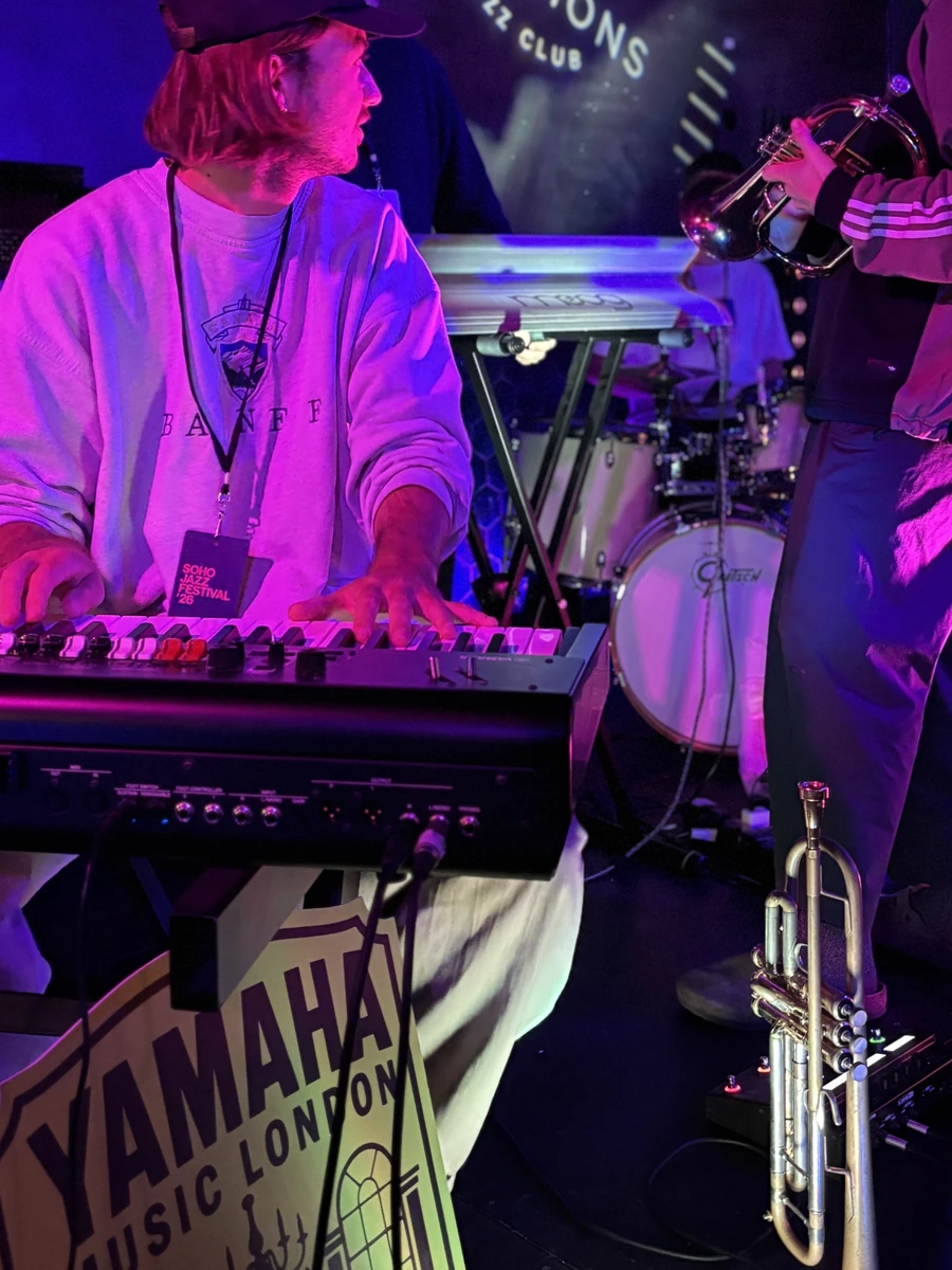 A keyboard player wearing a Soho Jazz Festival 26 pass, with a Yamaha Music London sign at the front of the stage and a trumpet waiting on its stand
