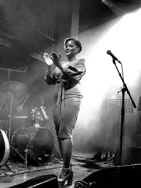 Riketté Genesis singing on stage, in black and white.